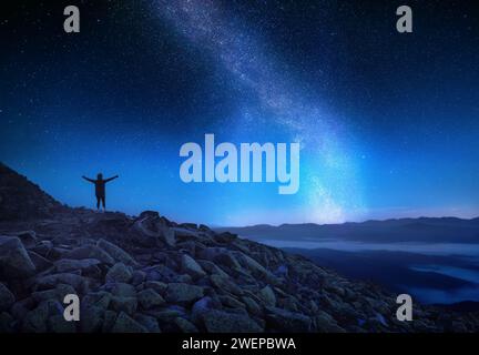 Glückliche Frau mit ausgestreckten Armen, die auf einem felsigen Hügel in den Bergen vor dem Sternenhimmel steht. Wunderschöne Nachtlandschaft mit heller Milchstraße. Bis Stockfoto