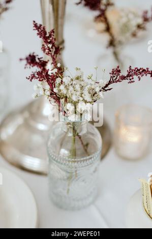 Blumen in einer Glasvase auf einer unberührten weißen Tischdecke, ergänzt durch flackernde Kerzen Stockfoto