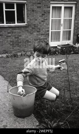 1960er Jahre, historisch, draußen in einem Garten, ein kleiner Junge, der Spaß beim Gartenarbeiten hat... Plastikeimer mit Wasser, kleine Gießkanne in der einen Hand und in der anderen eine Packung Ryders Samen Nemesia, England, Großbritannien. Stockfoto