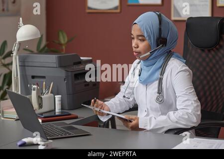 Junge Ärztin im Hijab, die Videoanruf mit ihrer Patientin hat und Dokumente ausfüllt Stockfoto