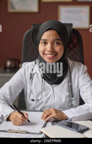Junge Ärztin im Hijab sitzt am Schreibtisch und lächelt in die Kamera, während sie Dokumente ausfüllt Stockfoto