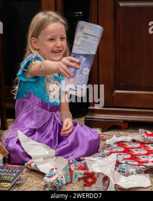 Das vierjährige Mädchen mit blonden blauen Augen sitzt auf dem Boden und zeigt ein Weihnachtsgeschenk, das sie geöffnet hat. USA. Stockfoto