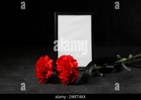 Leerer Trauerrahmen mit schwarzem Band und Nelkenblüten auf dunklem Hintergrund Stockfoto