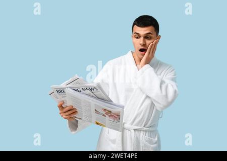 Porträt eines schockierten jungen Mannes im Bademantel mit Zeitung auf blauem Hintergrund Stockfoto