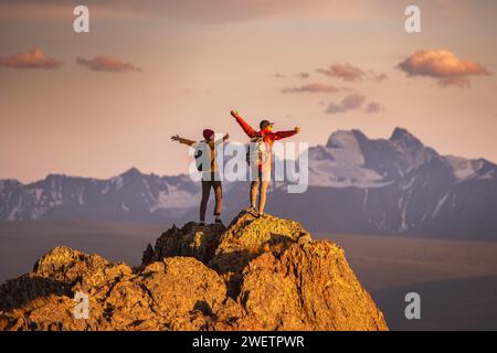 Zwei glückliche junge Touristen mit Rucksäcken stehen mit offenen Armen auf dem Gipfel des Berges und genießen den Sonnenuntergang Stockfoto