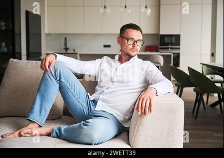 Charmanter Mann, der sich auf der Couch in seiner Wohnung entspannt. Gutaussehender junger Mann, der nachdenklich wegschaut. Mann in legeren Kleidern, der auf dem Sofa liegt, auf dem Hintergrund der modernen Küche. Stockfoto