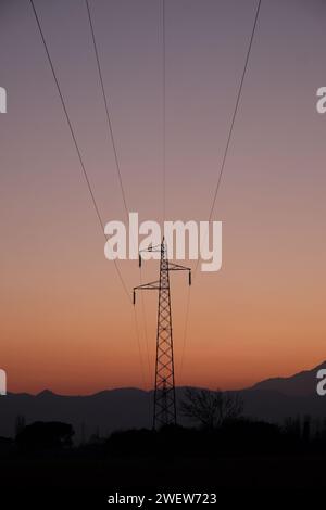 Die Silhouette eines Kraftübertragungsturms steht hoch in den beschaulichen Farbtönen eines Sonnenuntergangs, mit Stromleitungen, die sich bis zum Horizont erstrecken. Stockfoto