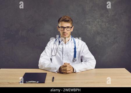 Porträt eines selbstbewussten ernsten jungen männlichen Gesundheitspersonals, der am Tisch auf grauem Hintergrund sitzt. Stockfoto