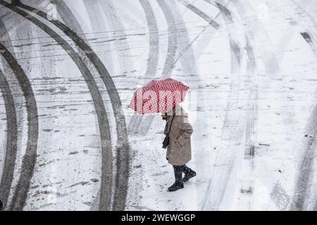 Frau mit einem rot-weiß gepunkteten Schirm überquert eine schneebedeckte Straße im Stadtzentrum, Schnee, Winter, Köln, Deutschland. Januar: 2024 Frau mit Stockfoto