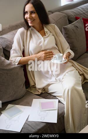Lächelnde schwangere Frau sitzt auf dem Sofa mit einer Tasse Kaffee Stockfoto