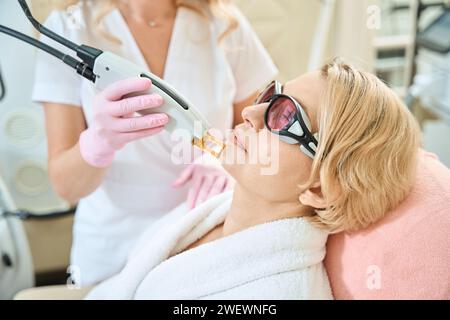 Beschnittene Kosmetikerin, die das Gesicht einer erwachsenen kaukasischen Frau mit Laser epiliert Stockfoto