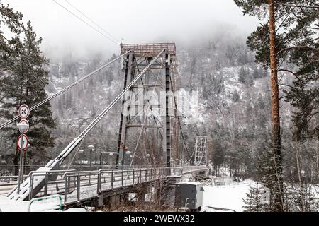 Metall-Hängebrücke für Autos und Fußgänger im Altai-Gebirge im Winter mit Schnee und Nebel auf den Bäumen ohne Menschen. Malerischer Platzgewinn Stockfoto