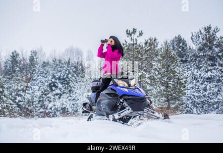 Mädchen auf dem Schneemobil im Wald. Fährt mit dem Schneemobil in den Bergen. Schneemobil im Schnee. Die Frau fährt mit dem Schneemobil. Reisen, Mädchen schaut durch Stockfoto