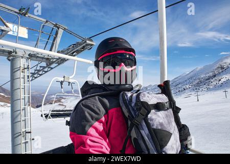 Der Mann geht den Skilift hoch. Person in Snowboardausrüstung: Stilvoller Schneehelm schwarz und rosa Farbe mit Schutzbrille, Rucksack. Seilbahnbau Stockfoto