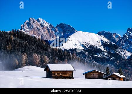 Hügeliges Ackerland mit Holzhütten und schneebedeckten Weiden auf der Seiser Alm, im Winter Gipfel der Geisgruppe in der Ferne. Kastelruth Stockfoto