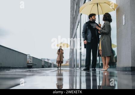 Zwei Geschäftsleute stehen unter einem gemeinsamen Schirm auf einem nassen städtischen Gehweg und diskutieren über die Arbeit mit einer anderen Person im Hintergrund. Stockfoto