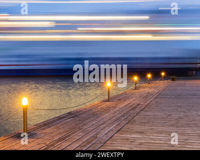 Dieses Bild erfasst die Bewegungsunschärfe eines Schiffes in einer Langzeitbelichtung, die von einem hölzernen Pier aufgenommen wurde, der während der blauen Stunde der Dämmerung mit Lichtern geschmückt ist. Stockfoto