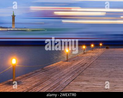 Dieses Bild erfasst die Bewegungsunschärfe eines Schiffes in einer Langzeitbelichtung, die von einem hölzernen Pier aufgenommen wurde, der während der blauen Stunde der Dämmerung mit Lichtern geschmückt ist. Stockfoto