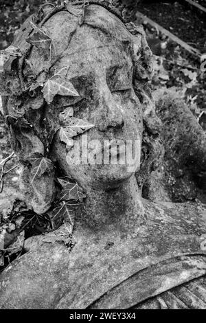 Gruselige alte Sandsteinstatue Frauengesicht mit einem Efeukranz auf dem Kopf. Schwarzweißbild. Winter. Friedhof. Konzept, Tod, Mortalität. Stockfoto