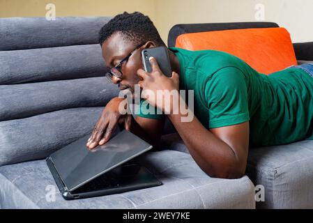 Junger Mann, der eine optische Brille trägt und zu Hause auf dem Sofa liegt und mit dem Handy spricht, während er den Laptop öffnet. Stockfoto