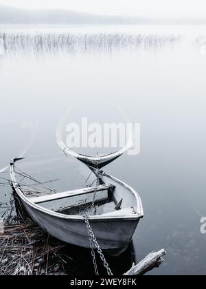 Zwei einsame Boote ruhen auf dem ruhigen Wasser eines Sees, umgeben von dem ruhigen Nebel eines frühen morgens. Stockfoto