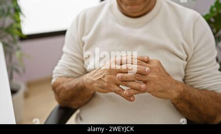 Ein hispanischer Mann mittleren Alters sitzt nachdenklich in einem Büro, die Hände mit einem verschwommenen Hintergrund in einem Innenraum zusammengeschlossen. Stockfoto