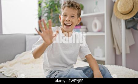 Entzückender blonder Junge voller Freude und Zuversicht, der zu Hause entspannt auf dem Sofa im Wohnzimmer sitzt, aber fröhlich mit der Hand hallo sagte und grinste Stockfoto