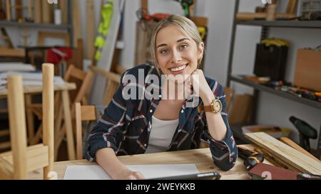 Lächelnde Frau mit blonden Haaren in einem karierten Hemd posiert in einer Tischlerei, umgeben von Holzwerkzeugen. Stockfoto