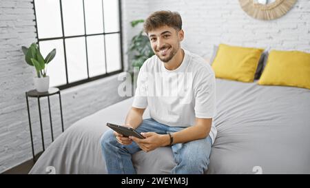 Selbstbewusster junger arabischer Mann, der seinen Morgen gerne genießt, sich bequem auf dem Bett entspannt und sich über das Touchpad in einem gemütlichen Schlafzimmer im Haus mit der Online-Welt beschäftigt Stockfoto