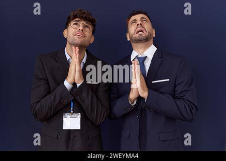 Zwei hispanische Geschäftsmänner, die Geschäftskleidung tragen, betteln und beten mit Händen, zusammen mit Hoffnungsausdruck im Gesicht, sehr emotional und besorgt. Be Stockfoto