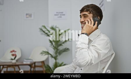 Ein selbstbewusster junger hispanischer Mann führt lächelnde Geschäftsgespräche, sitzt auf einem Stuhl im Wartezimmer und verwaltet Anrufe drinnen per Smartphone Stockfoto
