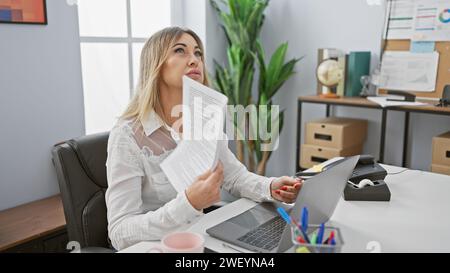 Reife weiße Frau fächert sich mit Papier in einem modernen Büroumfeld und blickt nachdenklich weg. Stockfoto