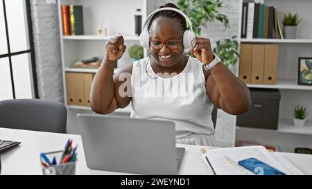 Feiertagszeit! Die afroamerikanische Chefin mit Zöpfen und Brillen rockt an ihrem Schreibtisch und gewinnt im Geschäft mit Laptops und Kopfhörern, die in offi arbeiten Stockfoto