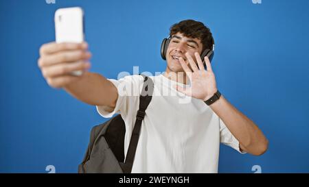 Cooler junger hispanischer Teenager, ein lächelnder Student, der in ein lebhaftes Videogespräch vertieft ist, isoliert vor einem blauen Hintergrund steht Stockfoto