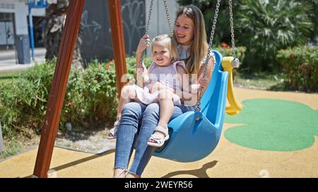 Kaukasische Mutter und Tochter, selbstbewusstes lächelndes Duo, das auf einer Spielplatzschaukel sitzt und fröhlich zusammen im Park spielt. Stockfoto