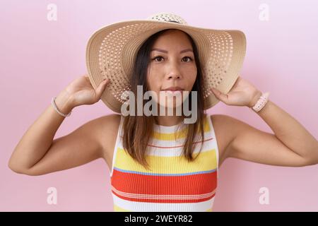 chinesische Frau mittleren Alters, die Sommerhut über rosa Hintergrund trägt, entspannt mit ernstem Gesichtsausdruck. Einfaches und natürliches Betrachten der Kamera. Stockfoto
