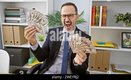 Fröhlicher Mann mittleren Alters mit grauen Haaren, erfolgreicher Geschäftsmann, grinsend, während er sein Vermögen im Büro zählt Stockfoto