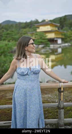 Fröhliche hispanische Frau mit Brille, die den goldenen Pavillon von kinkaku-JI bewundert. Ihr wunderschönes Lächeln strahlt, während sie die atemberaubende Aussicht in kyoto, japan, genießt. Stockfoto