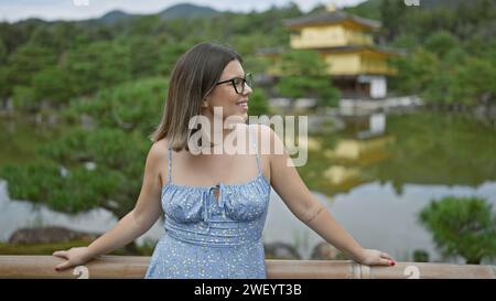 Fröhliche hispanische Frau mit Brille, die den goldenen Pavillon von kinkaku-JI bewundert. Ihr wunderschönes Lächeln strahlt, während sie die atemberaubende Aussicht in kyoto, japan, genießt. Stockfoto