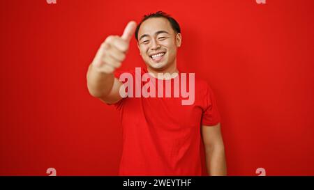 Selbstbewusster junger chinesischer Mann, der über rotem Hintergrund steht, den Daumen in einem „OK“-Schild nach oben blitzt, sein Lächeln strahlt Glück und coole Freizeitmode aus. Stockfoto