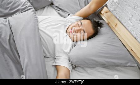 Junger, selbstbewusster chinesischer Mann genießt einen gemütlichen Morgenaufwachen, während er bequem in seinem Apartment-Schlafzimmer liegt und ein glückliches Lächeln auf seiner Hand spielt Stockfoto