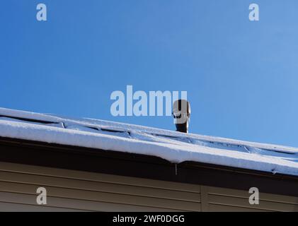 Ein Lüftungsblock mit Eiszapfen auf einem schneebedeckten Dach vor blauem Himmel. Stockfoto