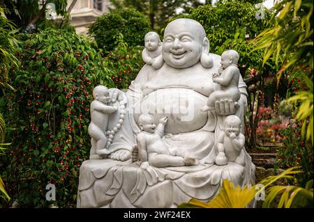 Herrliche weiße Marmorskulptur von Hotei, die in traditioneller Lotusposition sitzt und ein kleines Kind hält. Die heitere Statue ist von üppigem Grün und umgeben Stockfoto