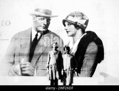 Zwei junge Mädchen posieren für einen Familienfoto vor einer Plakatwand aus den 1930er Jahren mit wohlhabenden Mann und Frau, CA. 1933. Stockfoto