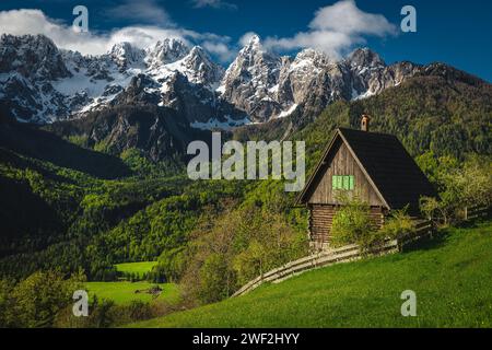 Majestätische Frühlingslandschaft mit gemütlicher Holzhütte auf der grünen Wiese und hohen schneebedeckten Bergen im Hintergrund, Dorf Srednji Vrh, Kranjska Gora, Ju Stockfoto