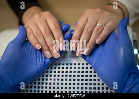 Nahaufnahme eines Nageltechnikers mit Handschuhen, der die Hände des Kunden hält, Seitenansicht der Hände des Nageltechnikers und des Kunden mit französischer Maniküre Stockfoto