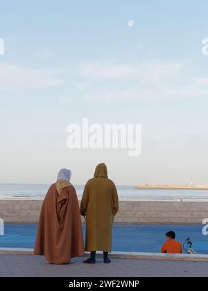 Einheimisches Paar in traditionellen Gewändern blickt auf den Atlantik mit dem Mond über n Essaouira, Marokko, 28. Januar 2024 Stockfoto