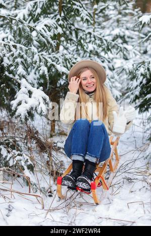 Schönes Mädchen, das auf einem Schlitten sitzt, ihren Hut hält und einen schönen schneebedeckten Wintertag genießt Stockfoto