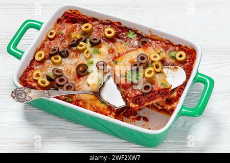 italienische Lasagne garniert mit Olivenringen und frischer Petersilie in grüner Auflaufform auf weißem Holztisch, Nahaufnahme Stockfoto