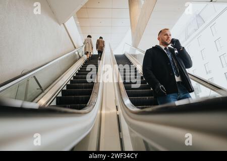 Ein stilvoller Geschäftsmann im Wintermantel führt während er eine Rolltreppe hinunterfährt, um die Mobilität und die Konnektivität in der Stadt darzustellen. Stockfoto